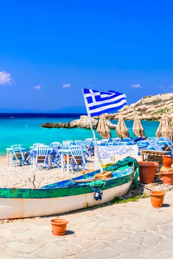 Strand met blauw-witte stoelen en parasols van stro. Een oud boot met de Griekse vlag staat op de voorgrond. Op de achtergrond turkooisblauw water en een rotsachtige kust bij een heldere lucht.