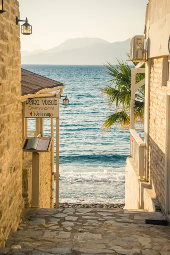 Een smal, geplaveid pad leidt tussen zandkleurige gebouwen met lantaarns naar de zee. Links hangt een bord "Petra Vassalo Restaurant Welcome", aan het einde staat een palmboom voor zachte golven.