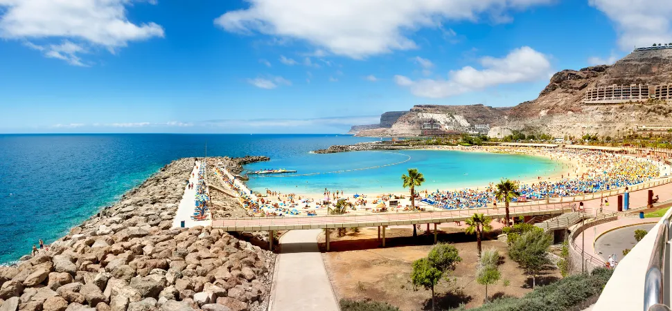 Panoramisch uitzicht op een zonnig strand op Gran Canaria, omlijst door helderblauw water en rotsachtige heuvels. Veel mensen relaxen onder parasols terwijl palmbomen het zandstrand omzomen.