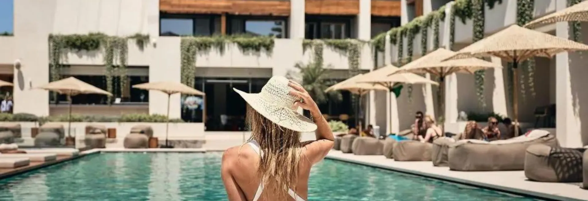 Een vrouw met een strooien hoed staat bij het zwembad en kijkt uit over een luxe resort met parasols en ligbedden. De ideale plek voor een all-inclusive vakantie in Griekenland.