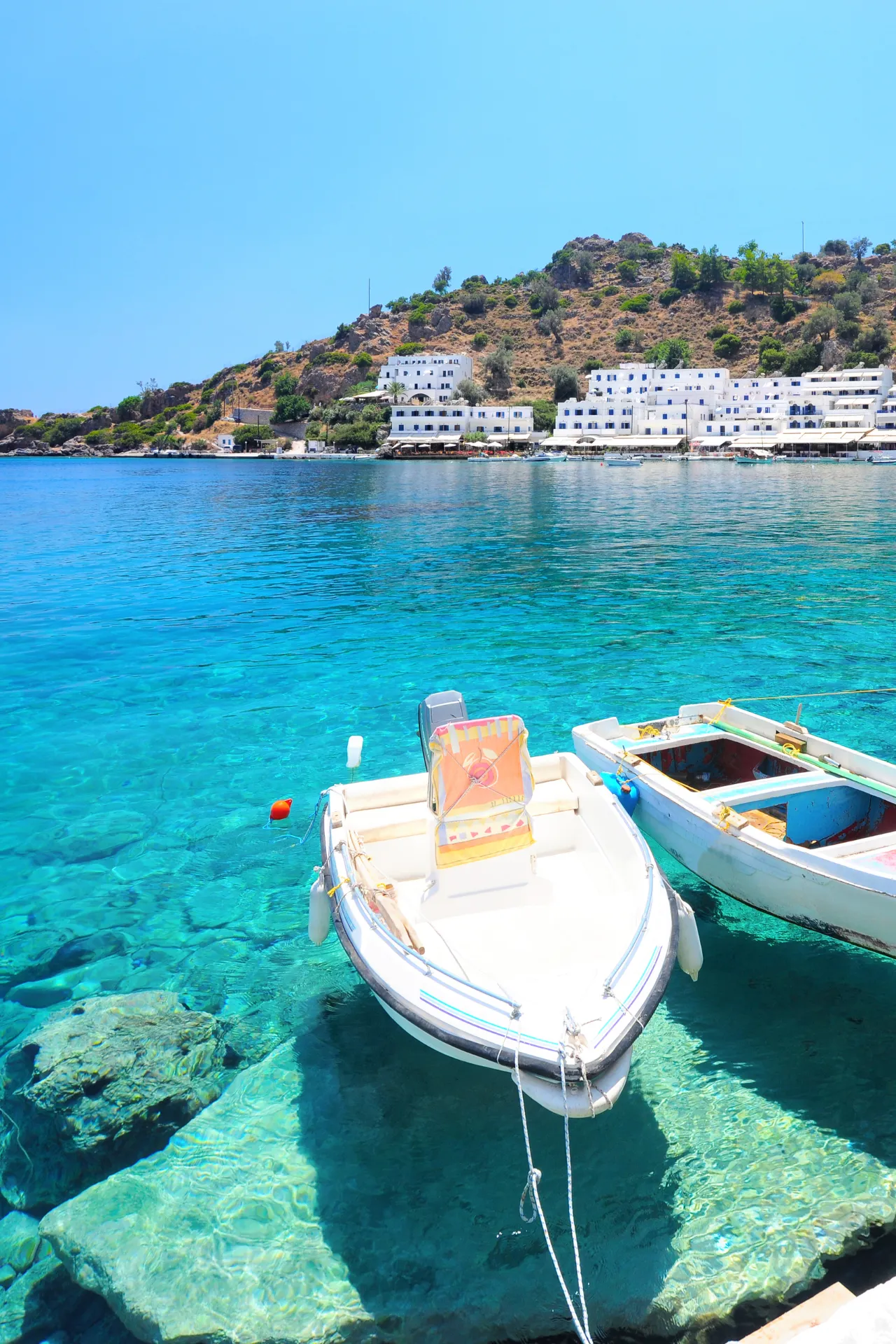 Twee boten dobberen op turquoise blauw water in een haven op Kreta. Op de achtergrond zijn witte gebouwen te zien tegen een groen heuvellandschap. Heldere zomerse sfeer.