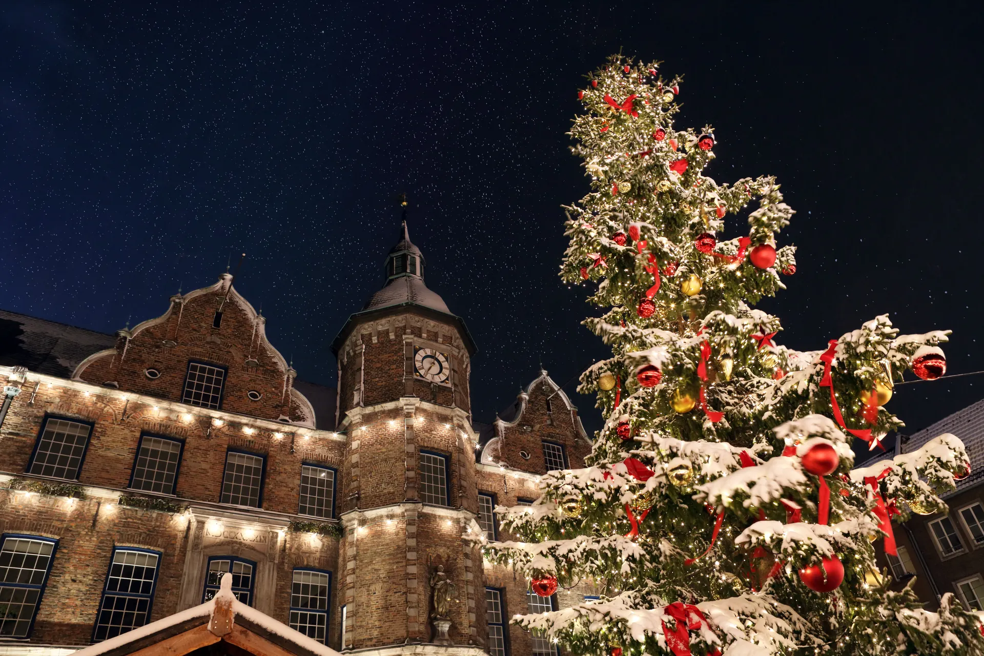 Besneeuwde kerstboom met rode linten voor een historisch gebouw met klok