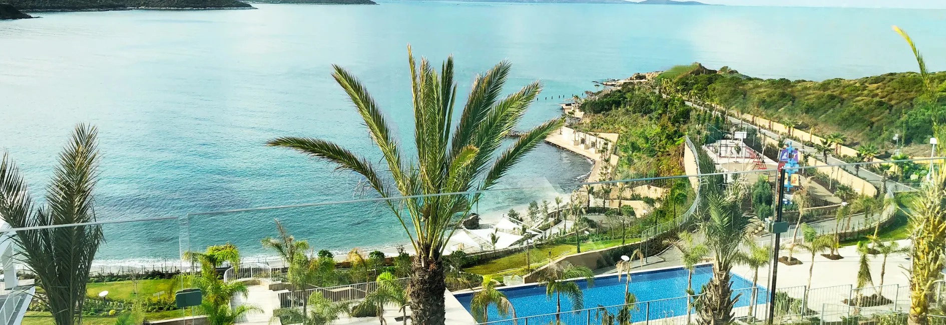 Uitzicht op een Turkse kustlijn met palmbomen op de voorgrond, een blauw zwembad en een strand met kristalhelder water. All Inclusive Turkije belooft ontspanning en luxe in deze idyllische omgeving.