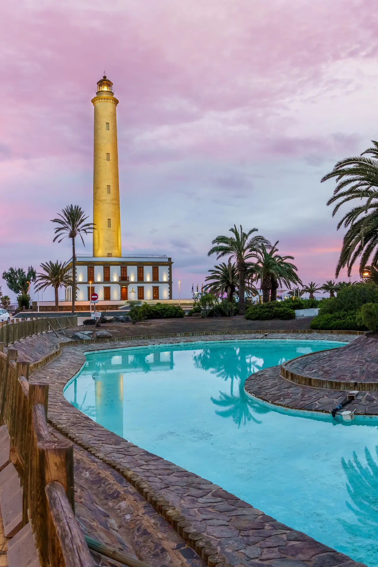 Een hoge, verlichte vuurtoren staat links voor een roze-blauwe lucht. Op de voorgrond slingert een gebogen zwembad, omringd door palmen en stenen paden.