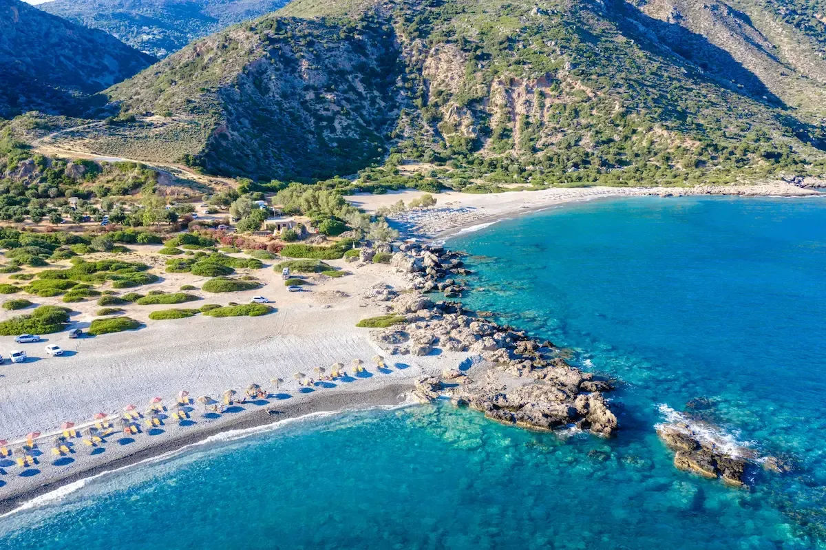 Bovenaanzicht over een strand met parasols en loungebedden aan een helderblauwe zee met in de achtergrond een berg met bomen erop