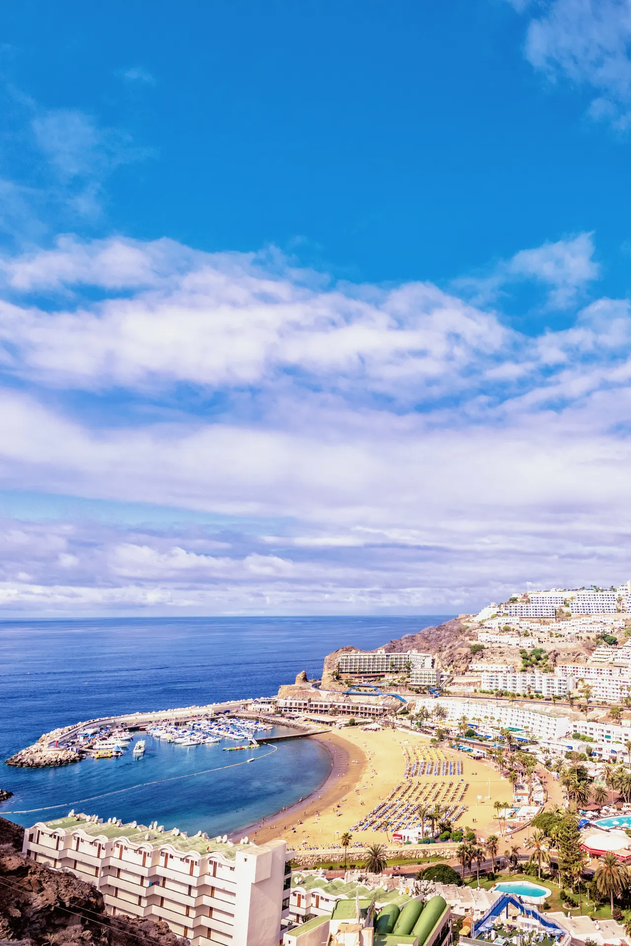 Luchtfoto van een kustlandschap met een baai vol boten, een gouden zandstrand, omringd door gebouwen en heuvels, op de achtergrond de eindeloze blauwe zee en een licht bewolkte lucht.