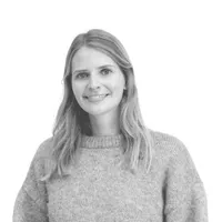 Een glimlachende vrouw met lang haar draagt een dikke, grijze trui. Ze kijkt recht in de camera, voor een witte achtergrond.