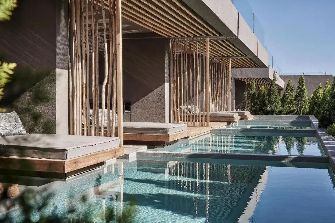 Luxe zwembadterrassen met houten ligstoelen en privacywanden onder moderne betonnen overkappingen. Kristalhelder water weerspiegelt de omgeving, omringd door groene planten rechts.