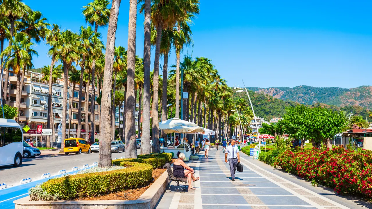 Promenade in Marmaris met palmbomen en bloembedden aan de rechterkant, rijbaan met auto's en gebouwen aan de linkerkant. Mensen wandelen en ontspannen op bankjes. Op de achtergrond groene bergen onder een heldere hemel.