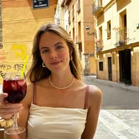 Jonge vrouw zit in een straatcafé en houdt een glas met een roodviolet drankje vast. Op de achtergrond oranje gebouwen met balkons en borden "Montaner Bakery" en "Carrer de Palomino".