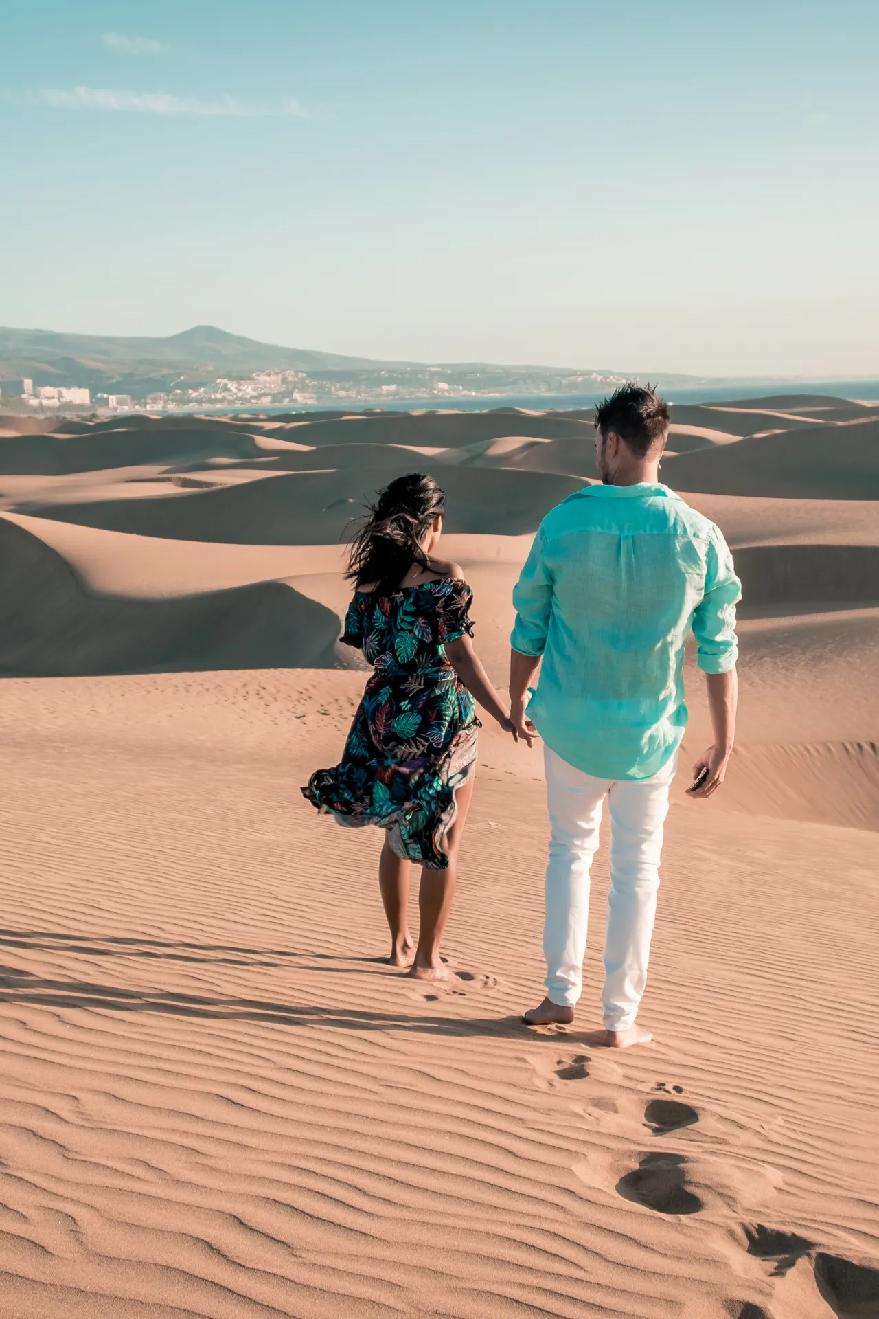 Een stel wandelt blootsvoets over een zandduin in een woestijnlandschap. De man draagt een turkoois overhemd, de vrouw een kleurrijke jurk. Op de achtergrond strekt de stad zich uit voor heuvels.
