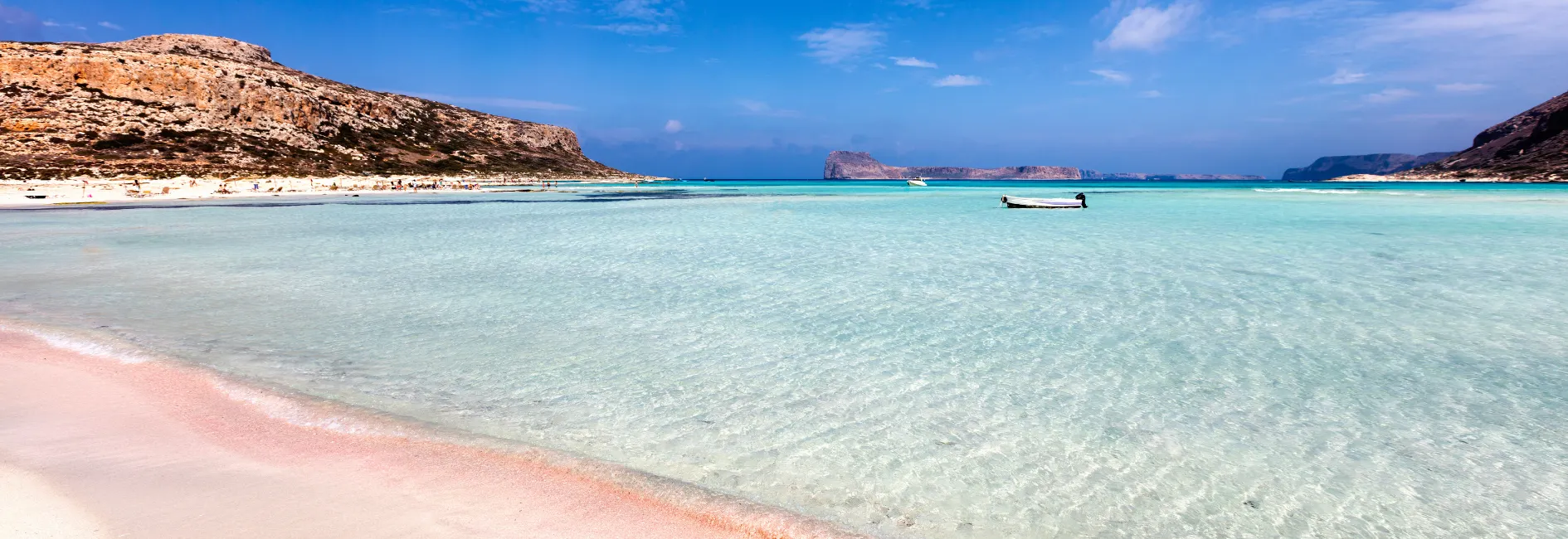 Strand met turkooisblauw water, fijn zand en kleine golven op de voorgrond. Rechts een boot in het water. Aan de horizon rotsachtige heuvels en een heldere, blauwe lucht. Kustlandschap in een ontspannen sfeer.