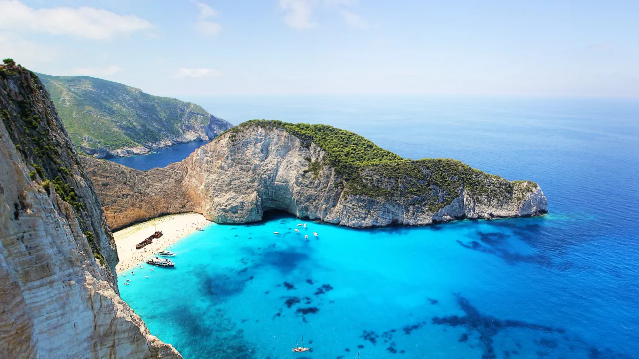 Helder, turkooisblauw water omringt een halfronde, hoge rotspunt met weelderig groen op Zakynthos. Eronder ligt een klein, wit zandstrand. 