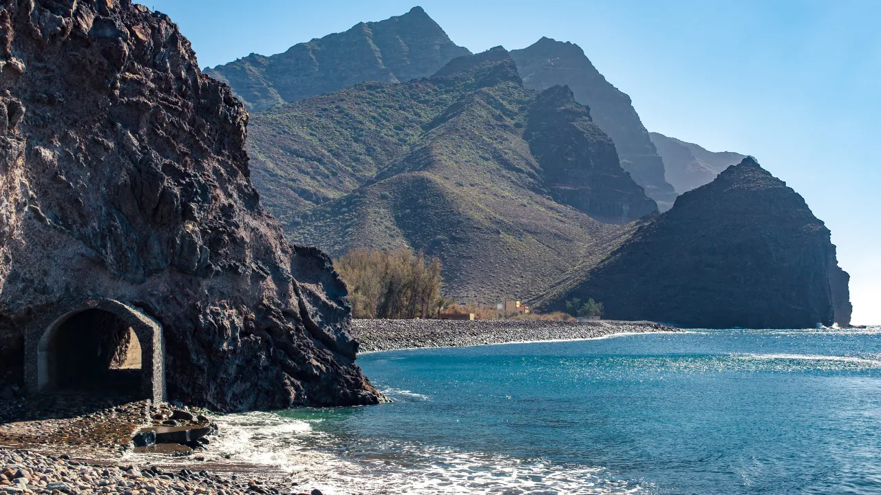 Steile kliffen en een tunnel aan de kustrand van Gran Canaria, met de zee aan de rechterkant en hoge bergen met kleine struiken aan de linkerkant. De lucht is zonnig