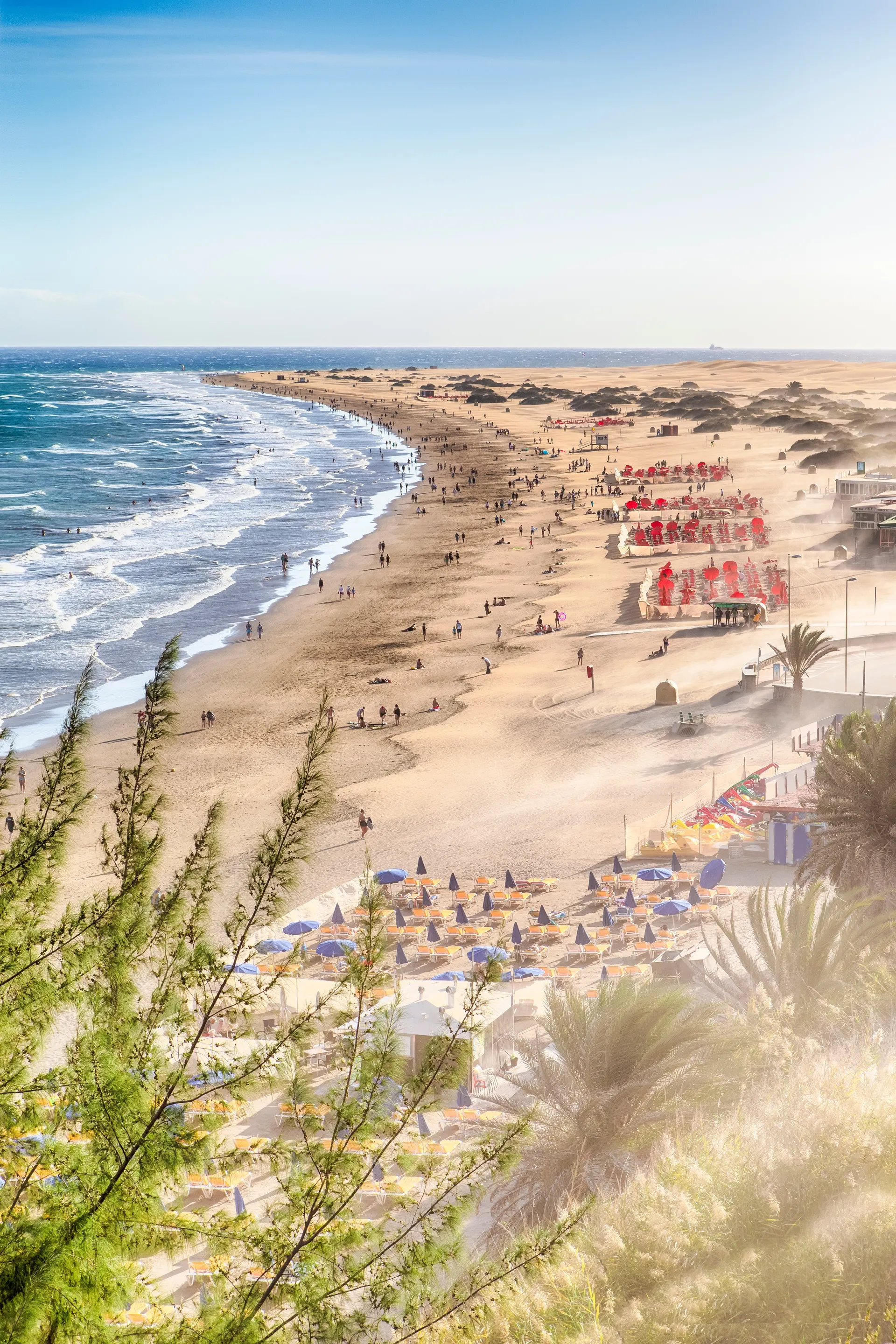 Uitgestrekt zandstrand met veel mensen die in het water en op het strand wandelen. Rechtsboven staan rode en blauwe parasols. Groene planten op de voorgrond, zee links met zachte golven.