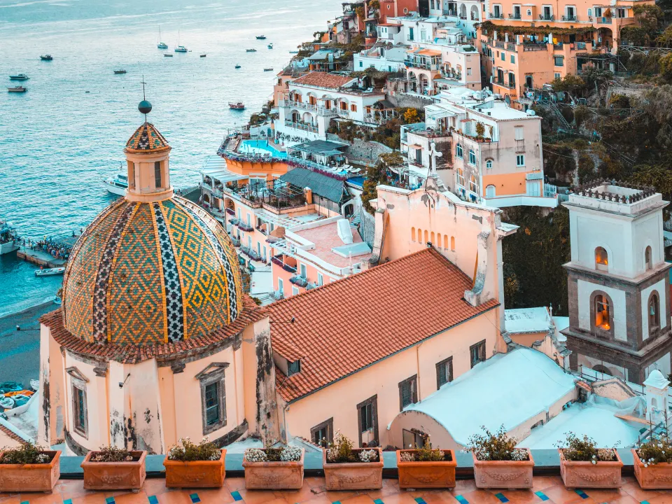 Koepel met kleurrijk mozaïek in het midden van de linkerhelft van het beeld, omringd door mediterrane gebouwen met terracotta daken. Rechts een klokkentoren, op de achtergrond de zee met kleine boten.