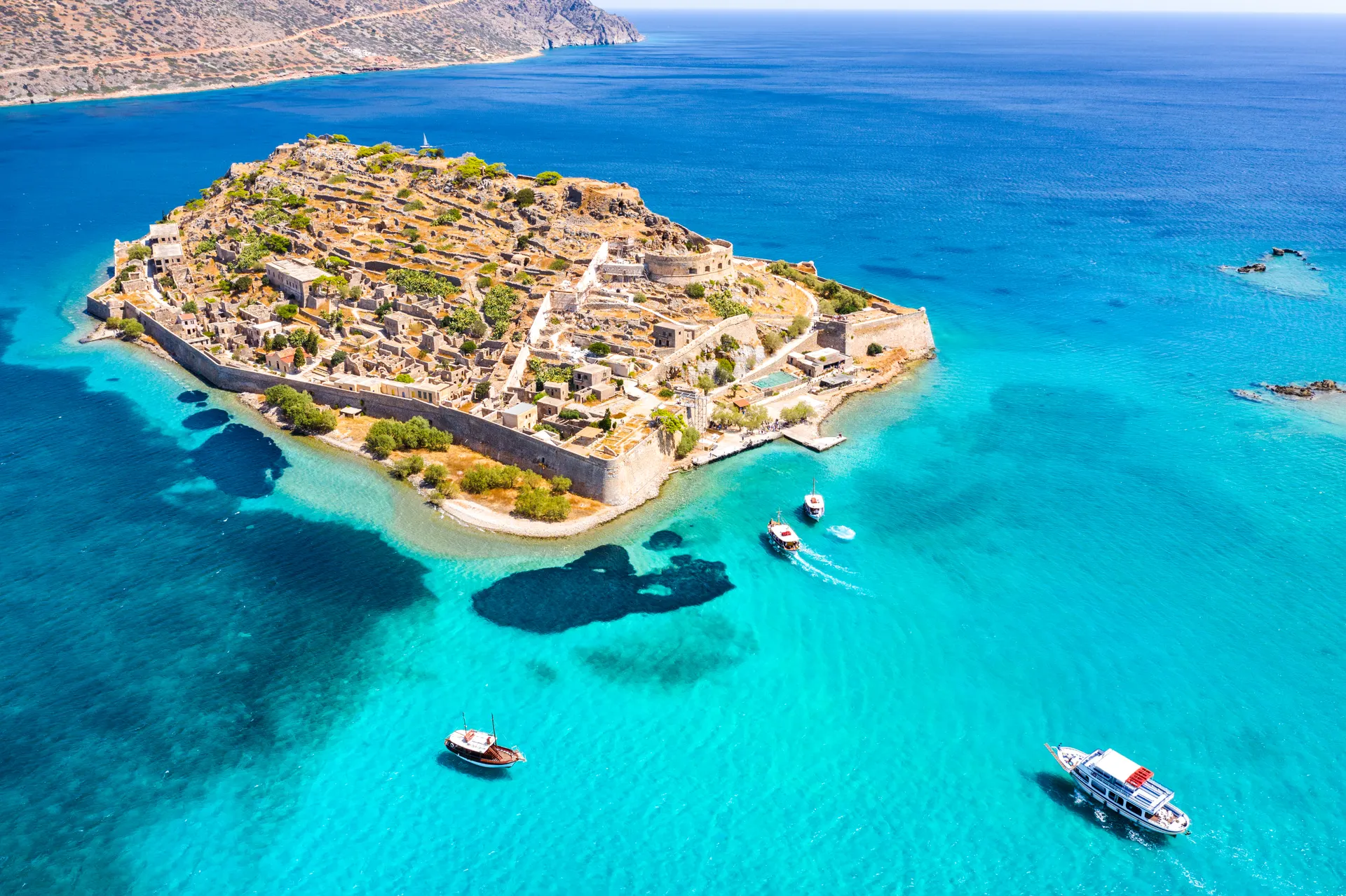 Het eiland Spinalonga vanuit vogelperspectief. Het eiland is omgeven door een muur en bebouwd met veel kleine huisjes. Omringd door de zee, met de kust van Kreta op de achtergrond