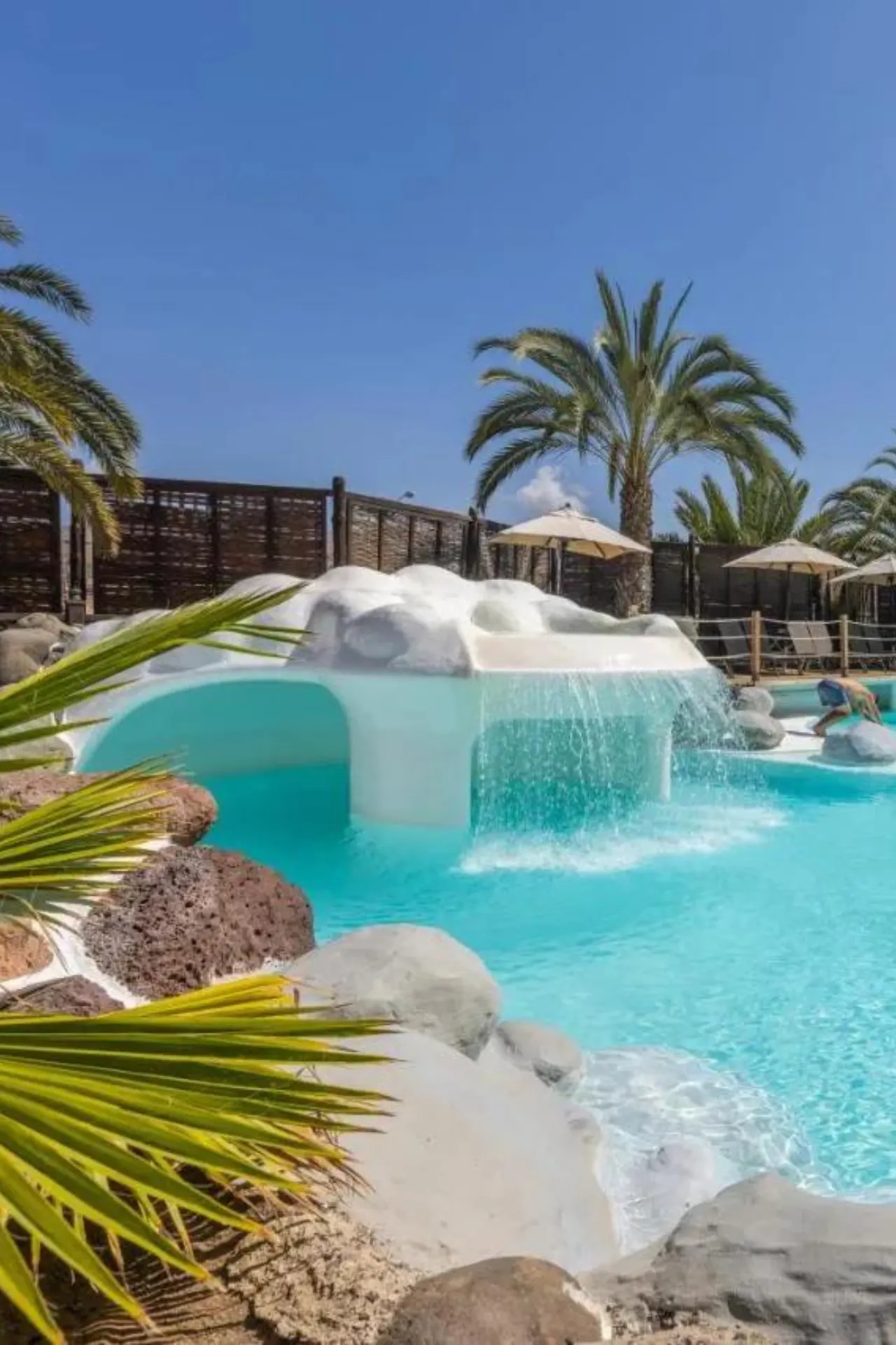 Turkooisblauw zwembad met rotsen en een palmboom op de voorgrond. Waterval stroomt over een witte boog. Op de achtergrond houten schuttingen, parasols en meer palmen onder een blauwe lucht.