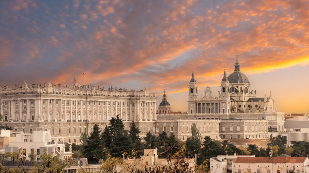 Paleis en kathedraal bij zonsondergang, links het paleis, rechts de kathedraal met koepels en torens, ervoor bomen en huizen, lucht in blauwtinten en oranje.