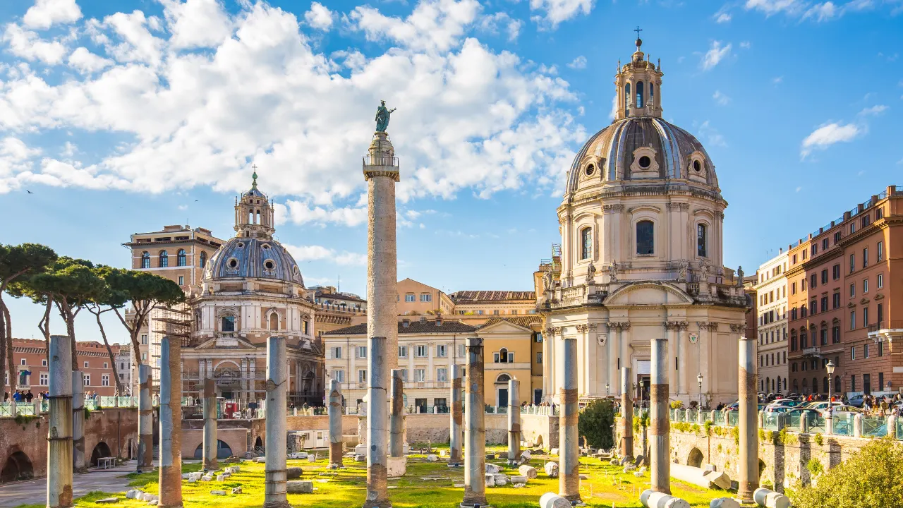 Historische gebouwen met koepels op de achtergrond, antieke zuilen op de voorgrond. Links staan bomen, rechts meerlagige gebouwen. Heldere lucht met enkele wolken erboven.