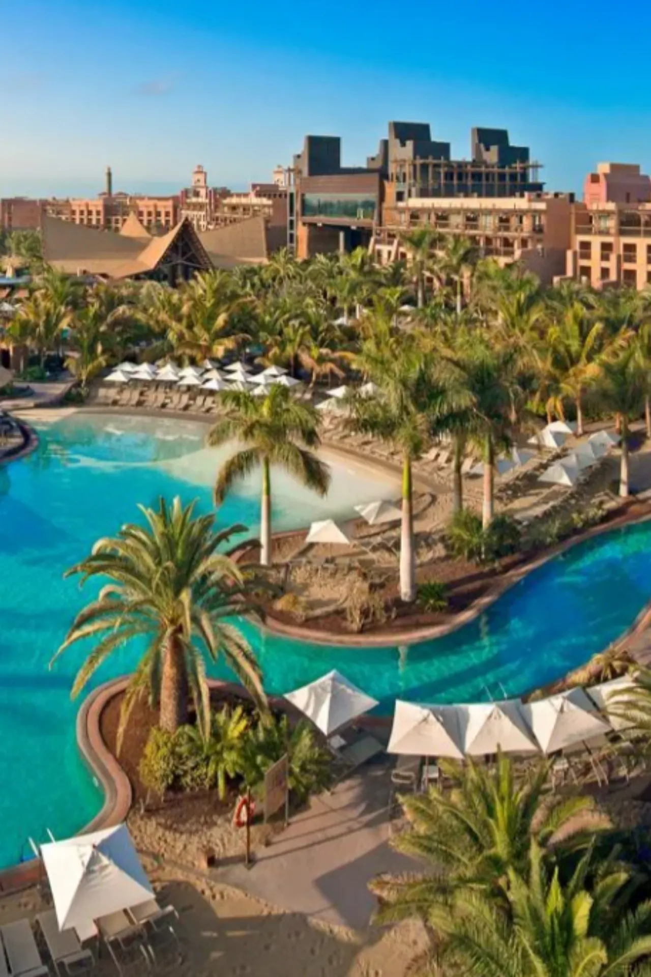 Luxe hotelcomplex met een zwembad in het midden, omgeven door palmen en ligstoelen onder witte parasols. Op de achtergrond moderne gebouwen met een blauwe lucht erboven.