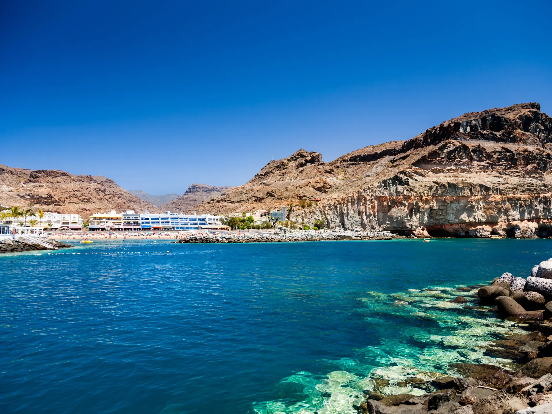 Heldere, turkooisblauwe zee op de voorgrond. De rotskust van Gran Canaria op de achtergrond, daarachter een pittoresk dorpje met witte gebouwen en palmbomen onder een blauwe hemel. Strand aan de linkerkant