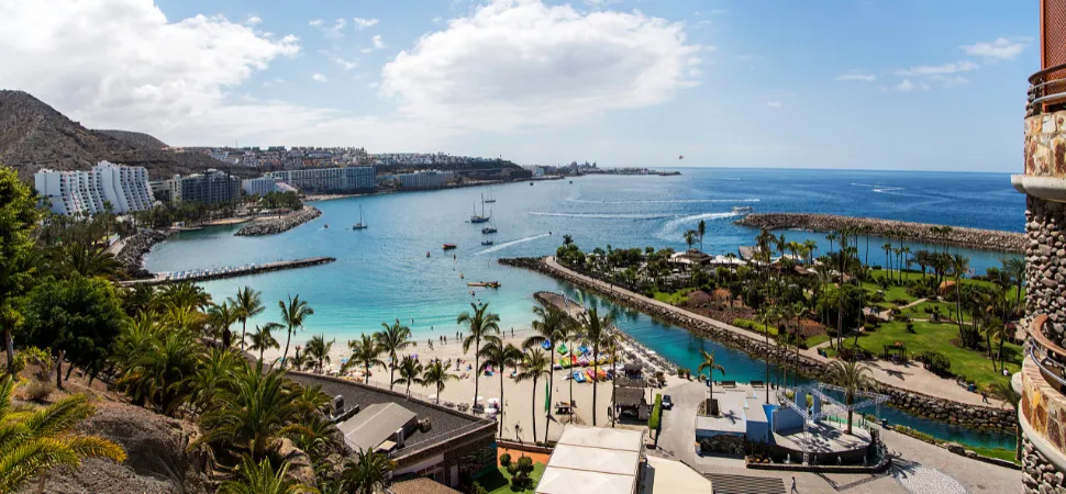 Panoramisch uitzicht op Gran Canaria: turkooisblauwe baai met zeilboten, lichtgekleurd zandstrand, omringd door palmbomen. Bergen en gebouwen op de achtergrond, waaronder een hotelcomplex. Rechts een ronde toren.